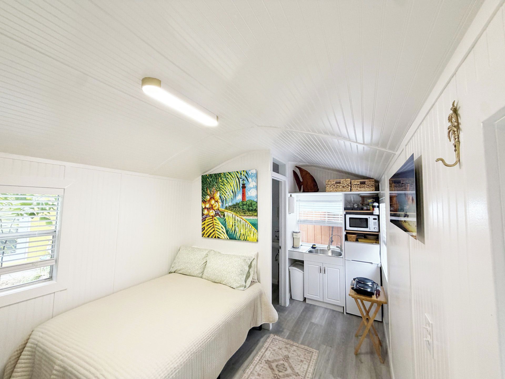 A compact room with a bed, a painting, a kitchenette, and a small window.
