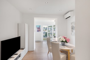 A modern living room with a flat-screen TV, a dining area with a table and chairs, and a view through a large window.