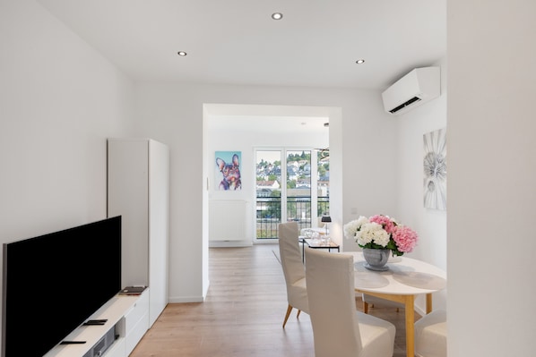 A modern living room with a flat-screen TV, a dining area with a table and chairs, and a view through a large window.