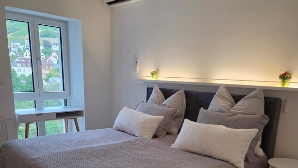 Ein Schlafzimmer mit einem großen Bett, einem Nachttisch, einem Fenster mit Blick auf Häuser und einer Wandleuchte.