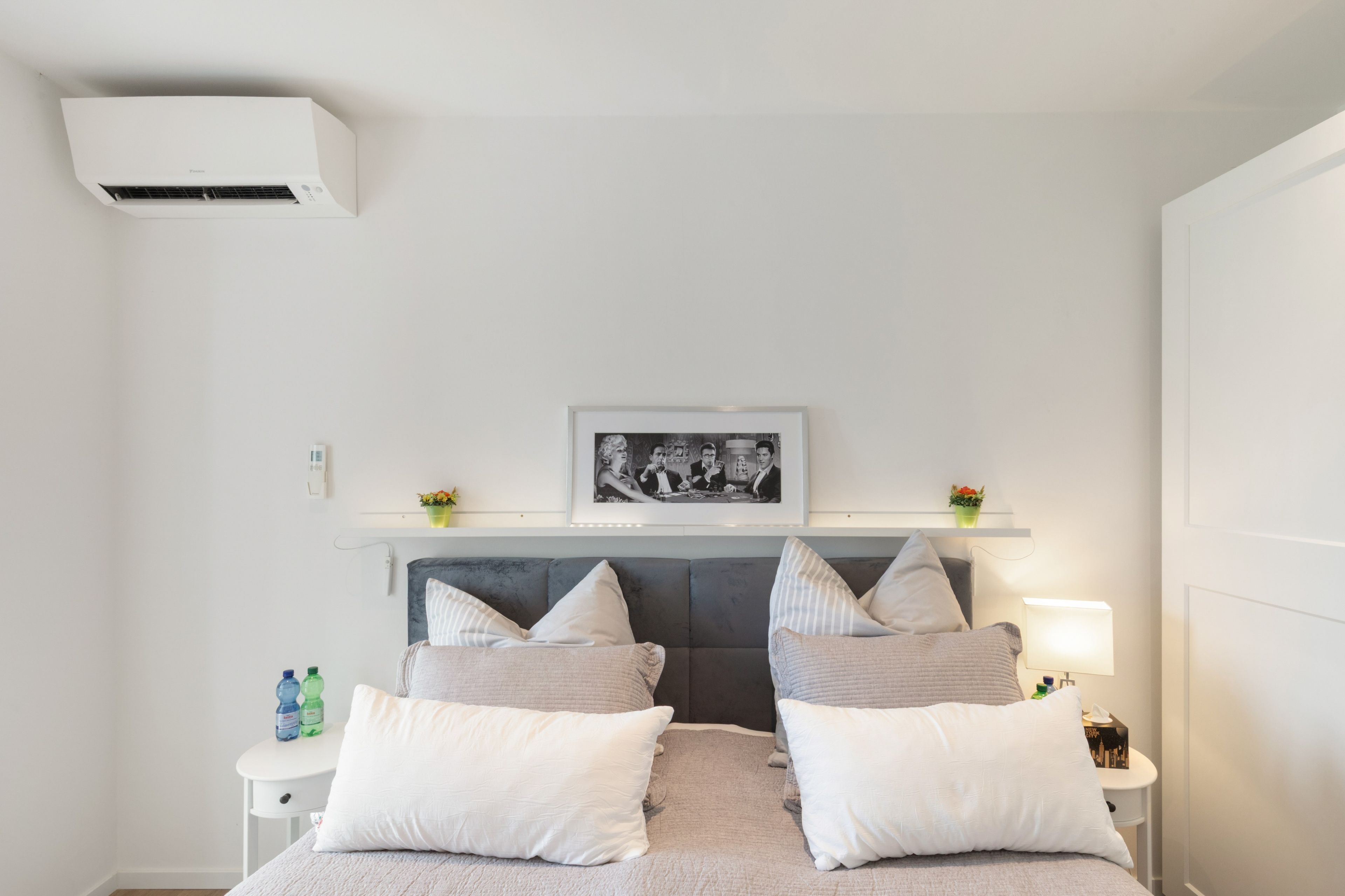 A modern bedroom with a bed, pillows, a bedside table, a nightstand, a framed picture, and an air conditioning unit.