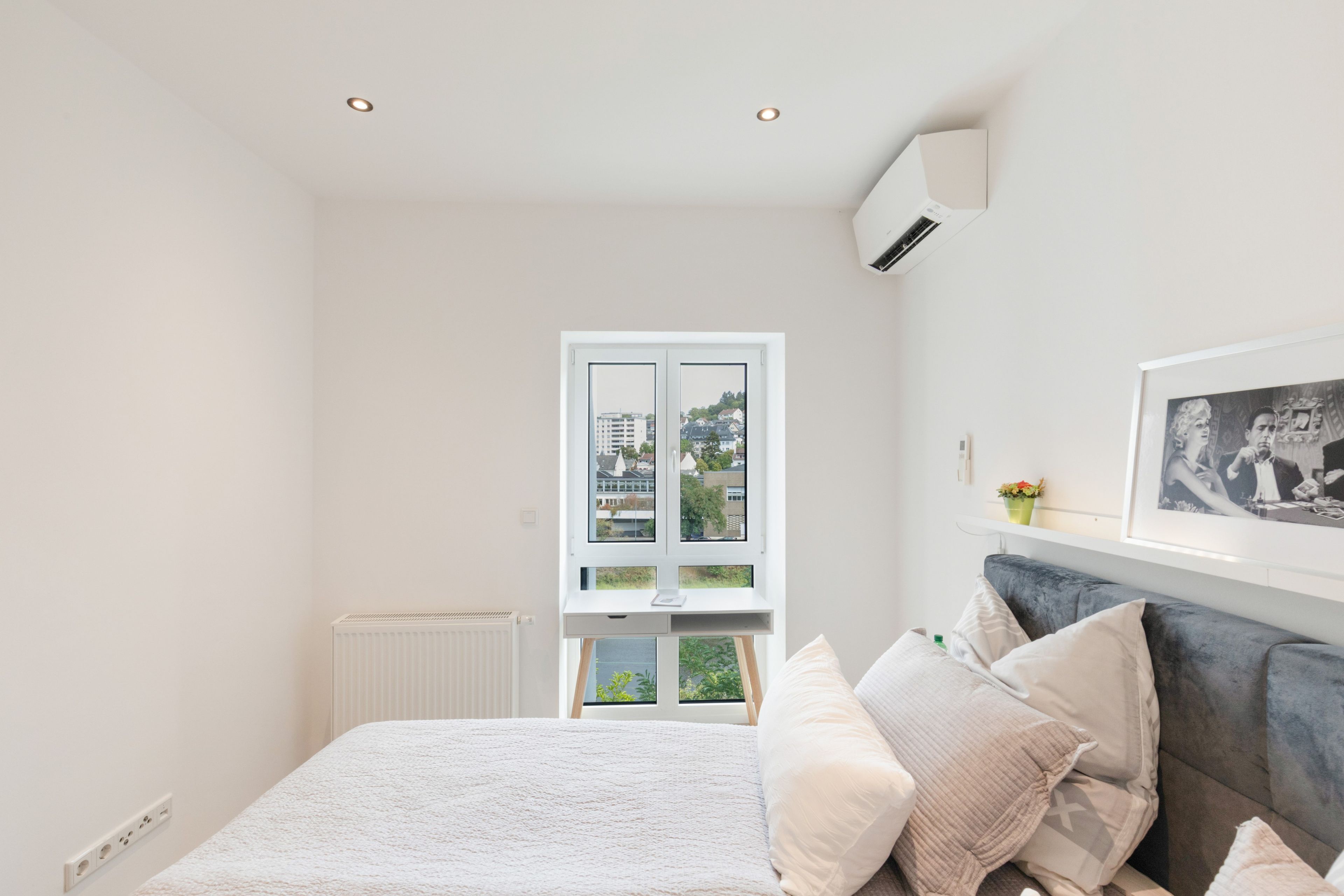 Ein Schlafzimmer mit einem Bett, einem Fenster mit Blick auf Gebäude, einem Heizkörper und einem gerahmten Bild an der Wand.