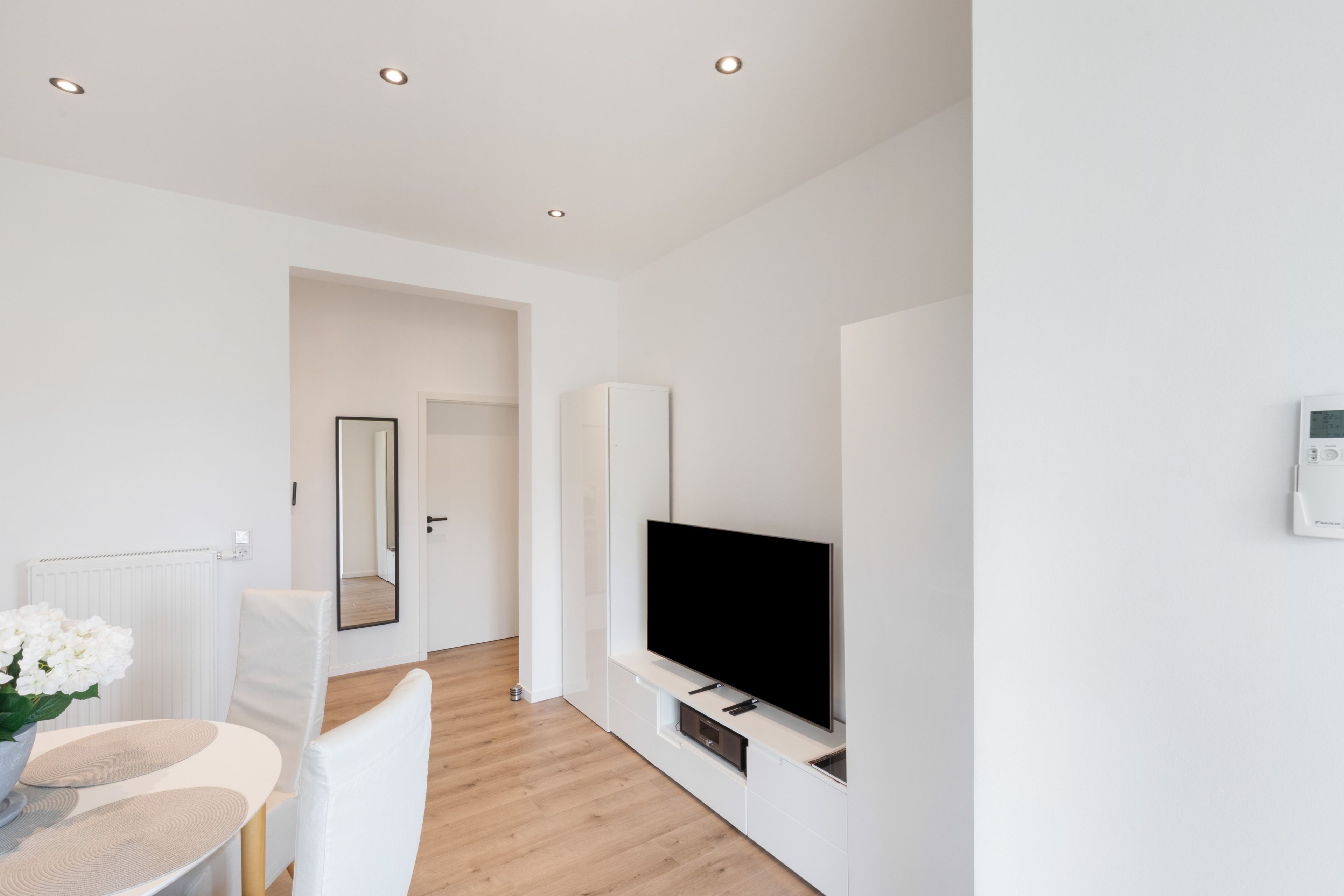 A modern, minimalistic living room with a flat-screen TV, a dining area with a table and chairs, and a hallway leading to another room.