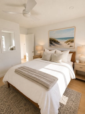 Een slaapkamer met een bed, bedlampen, een plafondventilator, een schilderij met een strandthema, en een spiegel.