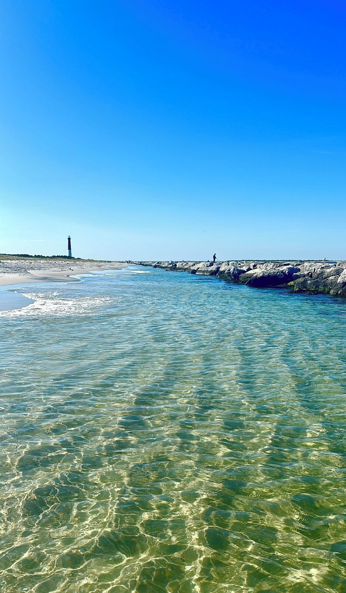 A coastal scene with clear water, a rocky breakwater, and a lighthouse in the distance.