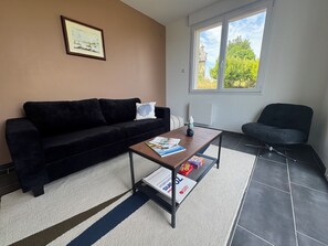 A living room with a black sofa, a coffee table with magazines, a framed picture on the wall, and a window with a view of trees. - House with ocean views near the GR34 and Royau Beach (Trévou-Tréguignec)