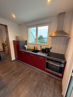 A modern kitchen with red cabinets, a stainless steel range hood, and a window overlooking a residential area. - House with ocean views near the GR34 and Royau Beach (Trévou-Tréguignec)