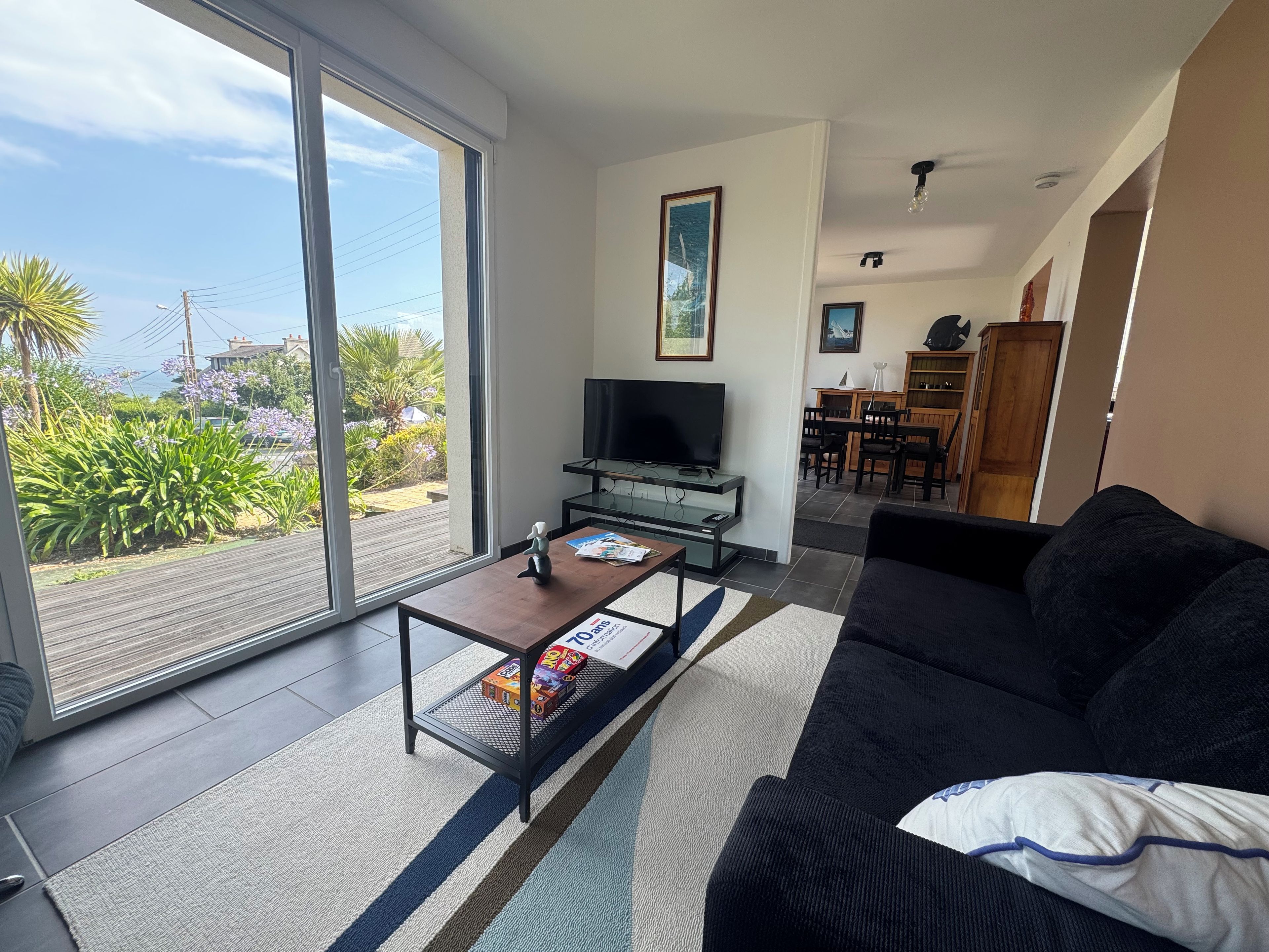 Sala de estar con sofá, una mesita de centro y televisor. Hay una puerta corrediza de vidrio que da a un balcón con plantas y vistas al exterior.