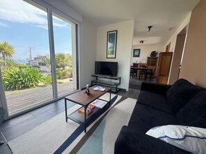Ein Wohnzimmer mit einer Couch, einem Couchtisch und einem Fernseher. Eine Glastür öffnet sich zu einem Balkon mit Pflanzen und Blick ins Freie.