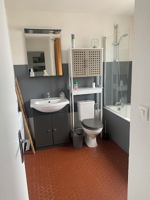 Et badeværelse med vask, toilet og bruser. Der er et spejl over vasken og en hylde med toiletartikler.