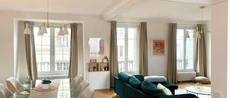 A modern living room with a dining area, large windows, and a view of the street.