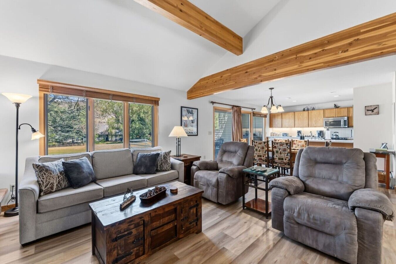 A spacious living room with a sofa, armchairs, and a coffee table. There is a kitchen in the background with wooden cabinets and a dining area.