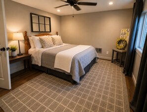 A bedroom with a bed, a nightstand, a lamp, a ceiling fan, a window with curtains, and a wall decoration.