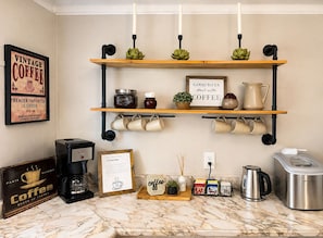 A kitchen with a coffee machine, a sign reading 'Good Days Start with Coffee,' and a shelf with mugs and a plant.