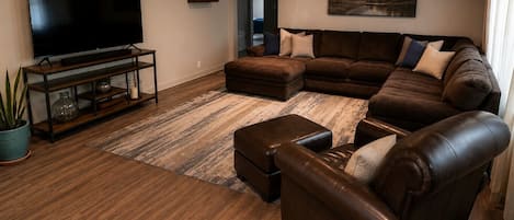A living room with a sectional sofa, a leather armchair, a flat-screen TV on a wooden stand, and a large painting on the wall.