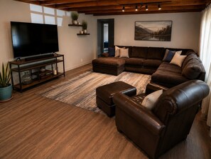A living room with a sectional sofa, a leather armchair, a flat-screen TV on a wooden stand, and a large painting on the wall.