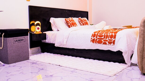 Cama com roupa de cama branca e almofadas laranja estampadas, cabeceira preta, tapete branco e um saco de roupa suja no chão.