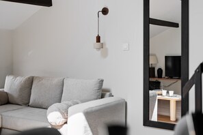 Ein modernes Wohnzimmer mit einem grauen Sofa, einem kleinen runden Tisch und einem Spiegel, der einen Fernseher und eine Lampe reflektiert.