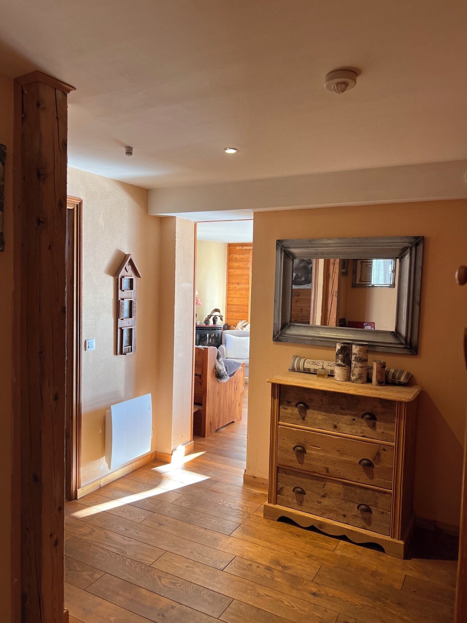 Un pasillo con suelo de madera, un mueble de madera con cajones, un espejo en la pared y una puerta que da a otra habitación.