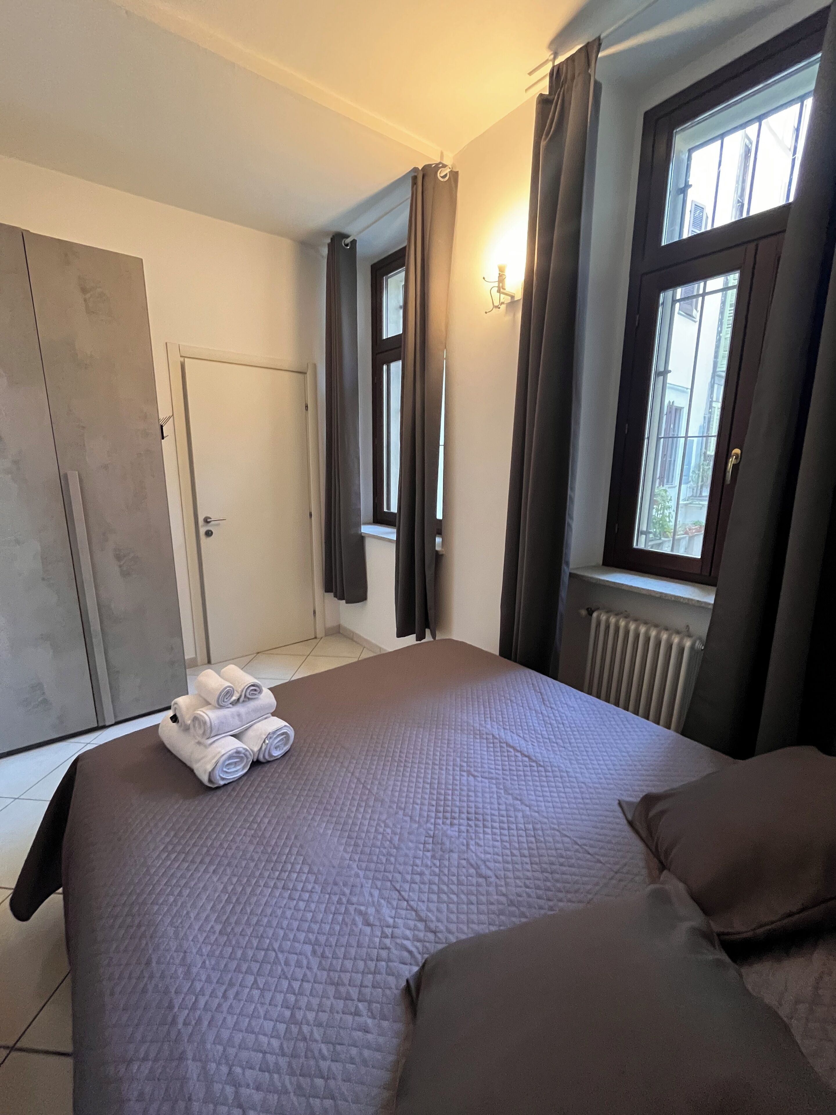 A bedroom with a bed, a radiator, a window with curtains, and a door.