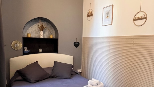 A neatly made bed with pillows and rolled towels, a wall-mounted shelf with decorative items, and a framed picture on the wall.