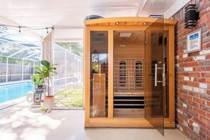 A glass-enclosed sauna with a wooden exterior, a pool visible outside, and a brick wall with a sign. - Enclosed Pool & Sauna Retreat Near Beaches (Fort Walton Beach)