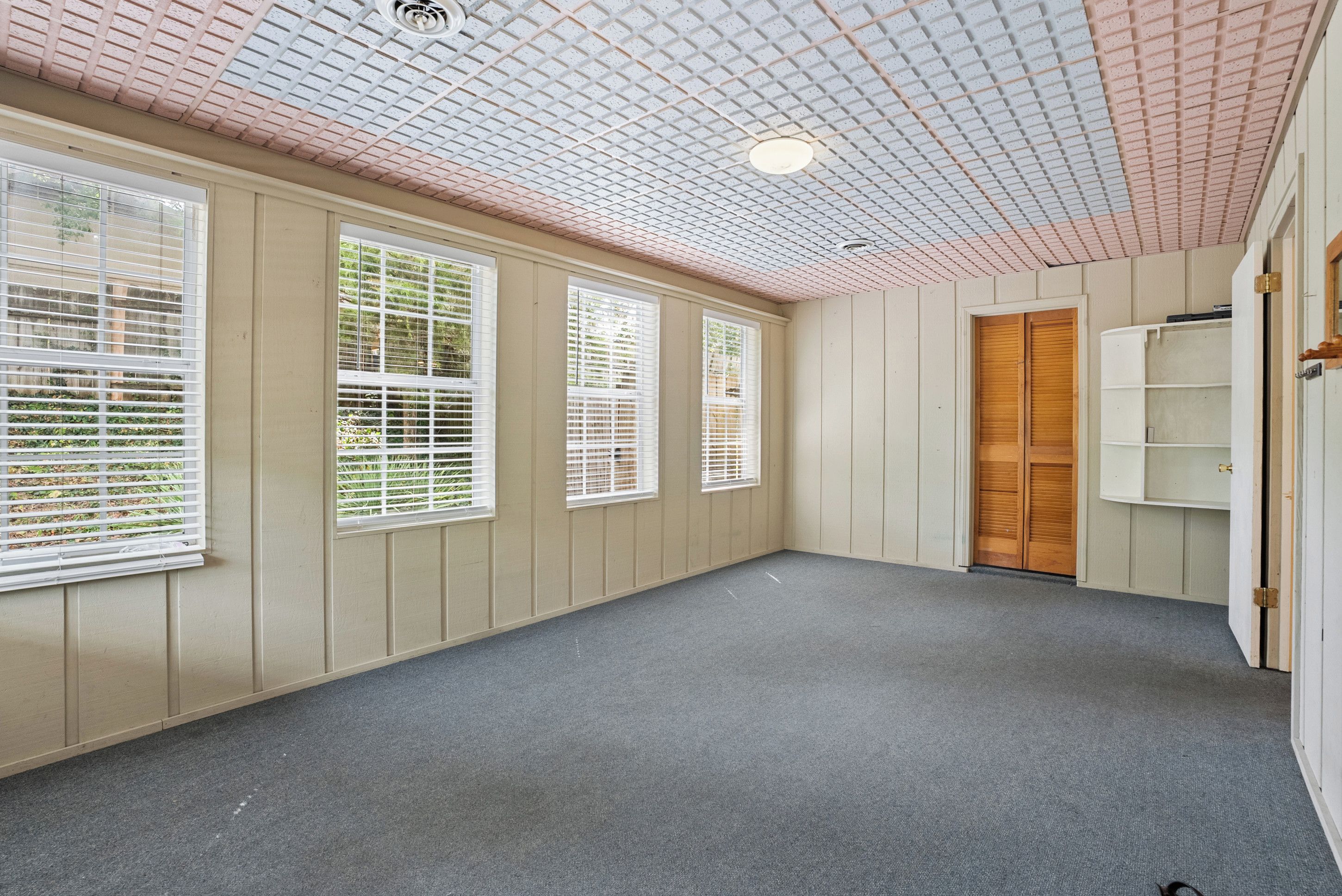 Une pièce avec un plafond en pente, de grandes fenêtres équipées de stores, une porte en bois et un plancher recouvert de moquette.