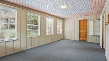 Une pièce avec un plafond en pente, de grandes fenêtres équipées de stores, une porte en bois et un plancher recouvert de moquette.