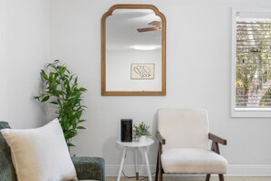 Ein Zimmer mit einem großen Spiegel, einem gerahmten Bild, einer Zimmerpflanze, einem kleinen Tisch mit einem Buch und einem Fenster mit Jalousien.