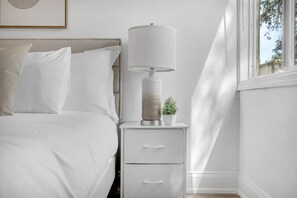 Ein ordentlich bezogenes Bett mit weißen Leinen und Kissen, ein Nachttisch mit Lampe, ein kleiner Topfpflanze und ein gerahmtes Bild an der Wand.