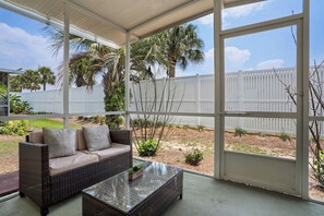 Un patio couvert avec un canapé en osier et une table basse en verre, une clôture blanche et des palmiers en arrière-plan.