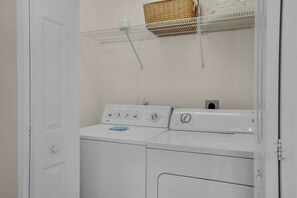 Une buanderie équipée de deux machines à laver et deux sèche-linge superposés, d’une étagère en métal avec un panier, et d’une porte blanche.
