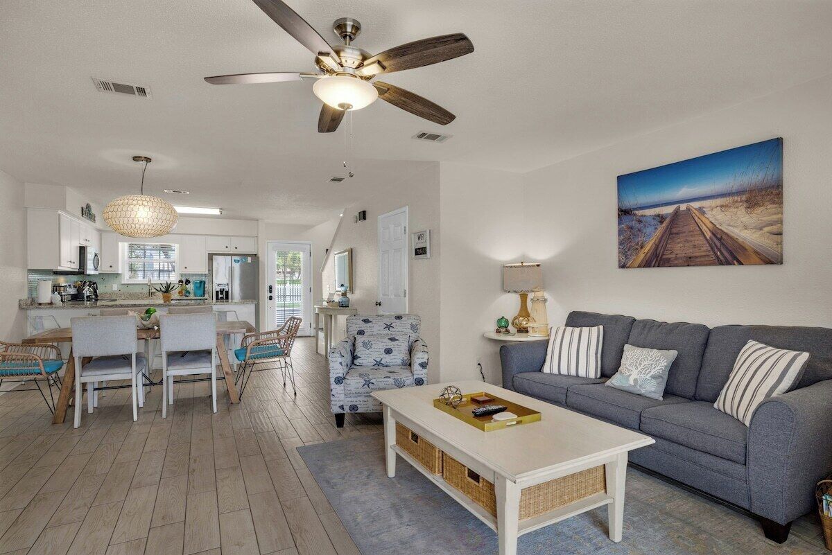 A spacious living area with a ceiling fan, a gray sofa with striped pillows, a dining table with chairs, and a kitchen in the background.