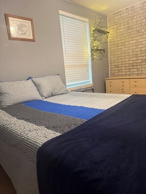 A bedroom with a bed, a dresser, a window with blinds, and a wall-mounted light fixture.
