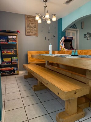 A dining area with a wooden bench and table, a bookshelf with board games, and a wall clock.