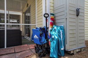 A covered porch with a wheelchair, a blue bag, and a teal bag leaning against a building. - Beachfront Townhome w/ Pool (Destin)