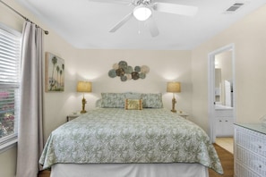A bedroom with a bed, two bedside lamps, a ceiling fan, a window with curtains, and a wall with decorative items. - Beachfront Townhome w/ Pool (Destin)