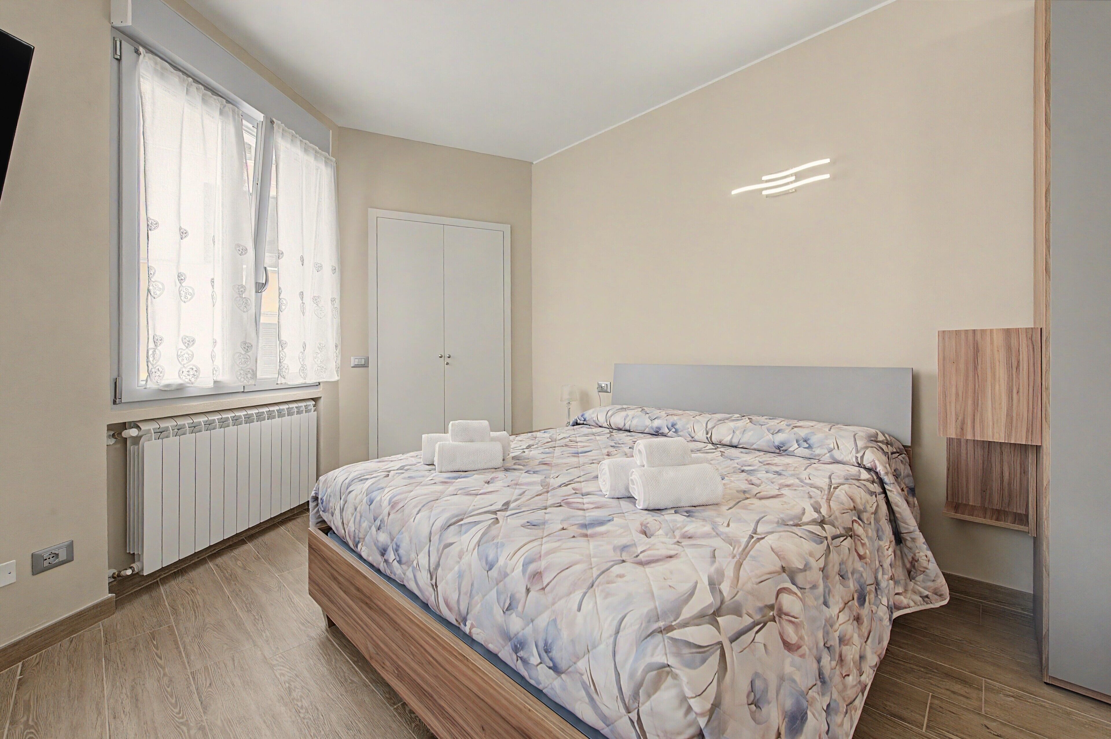 A bedroom with a bed, window, radiator, and a bedside table.