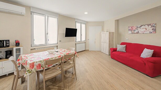 A compact living space with a dining area, a red sofa, a flat-screen TV mounted on the wall, and a radiator under the windows.