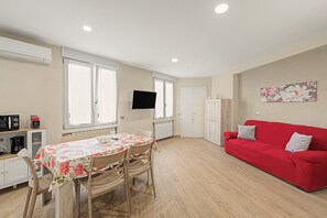 A compact living space with a dining area, a red sofa, a flat-screen TV mounted on the wall, and a radiator under the windows.