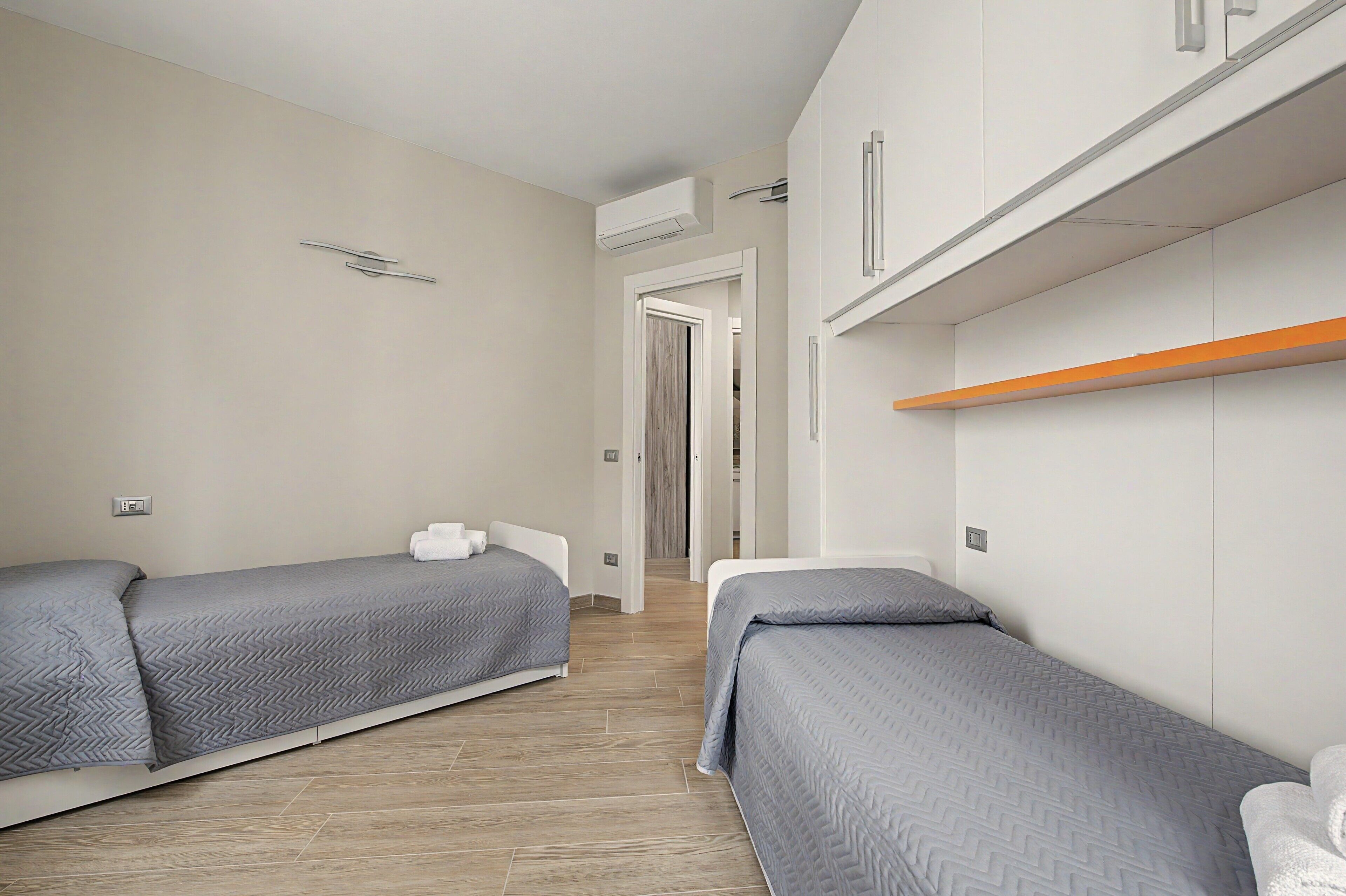 Habitación de hotel con dos camas individuales, piso de madera, un armario blanco y un estante pequeño.