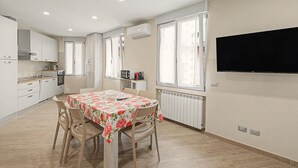 A modern kitchen with white cabinets, a dining table with floral tablecloth, and a large flat-screen TV mounted on the wall.