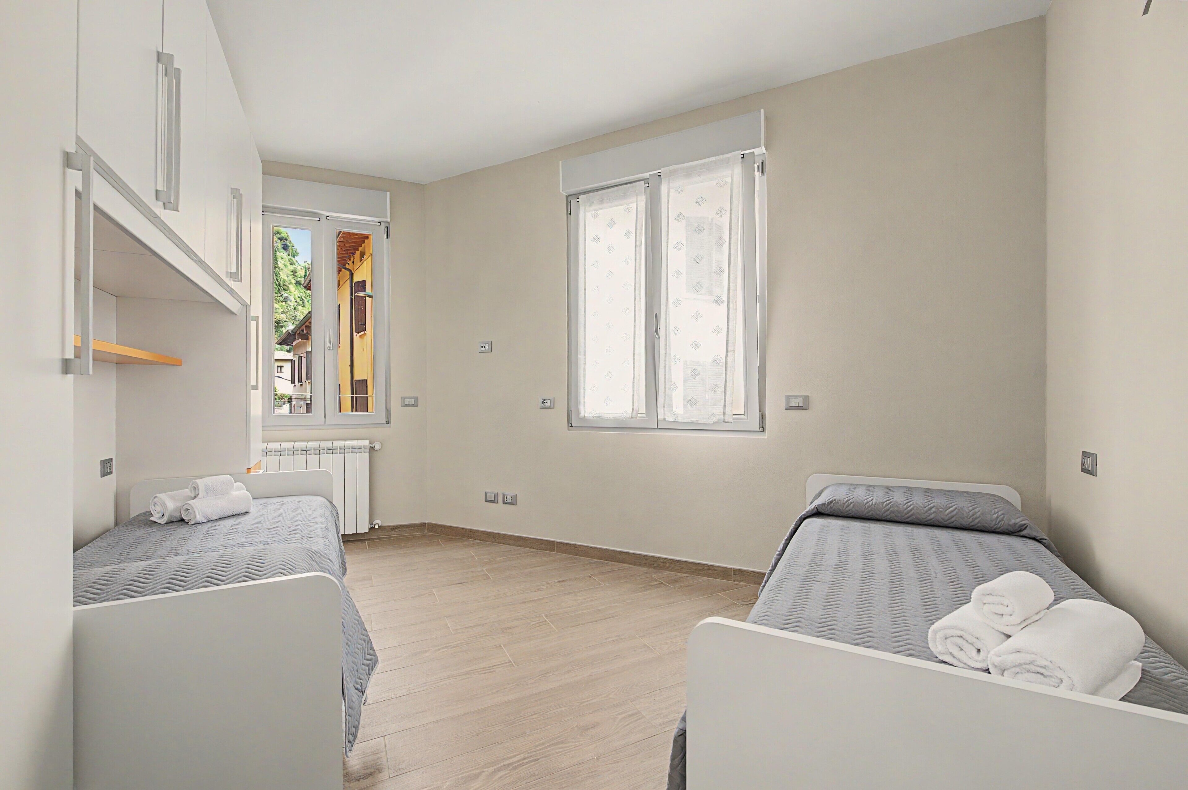 A hotel room with two beds, a window with curtains, a radiator, and a view of a building outside.