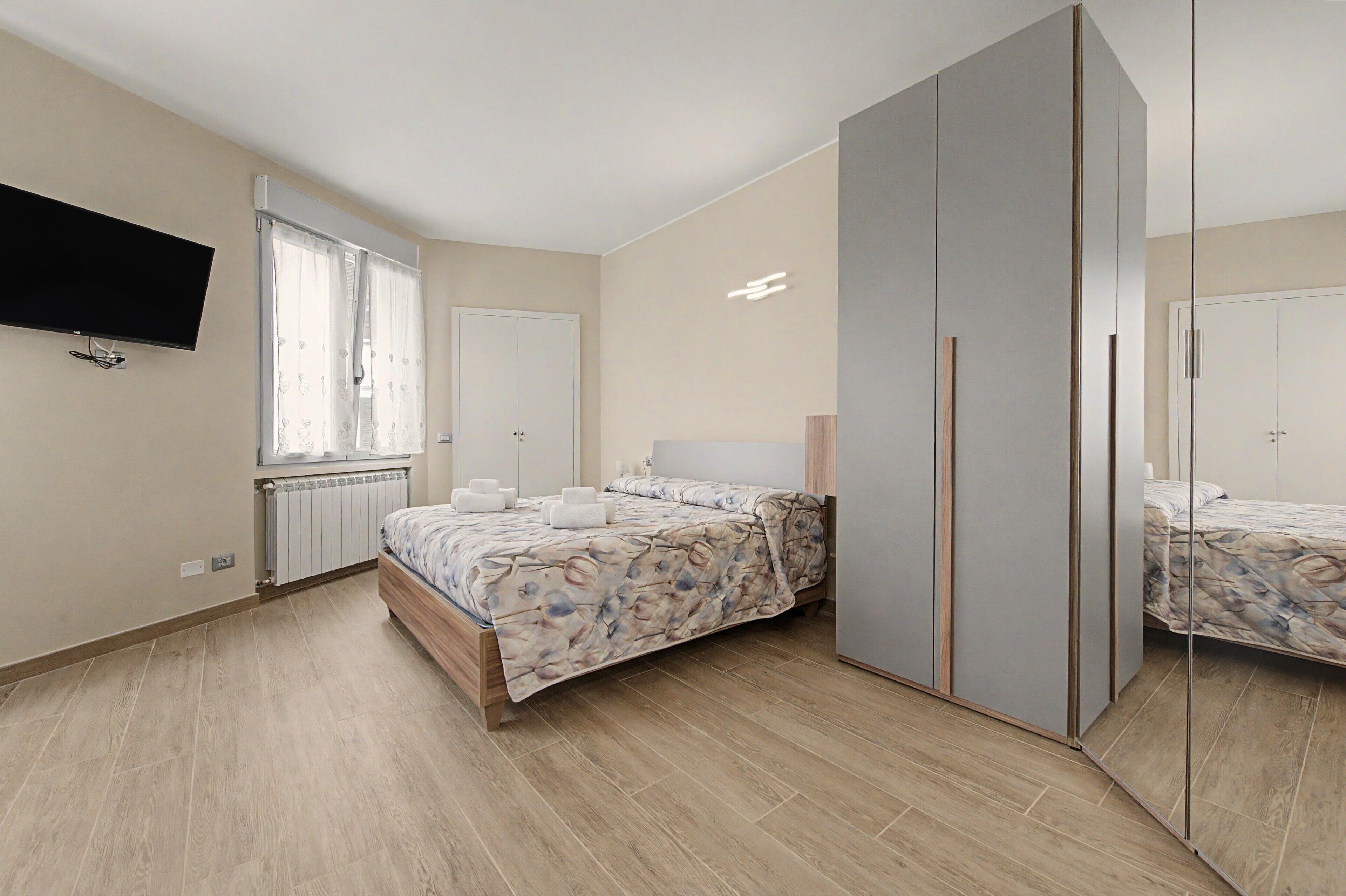 Un dormitori amb un llit, un armari, un mirall, una televisió i una finestra amb cortines.