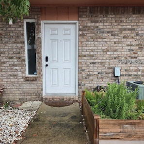 A white door with a small window above it, a brick wall, and a garden area with plants and a wooden planter. - Pet-Friendly Townhome w/ Fire Pit, 5mins to Beach (Fort Walton Beach)