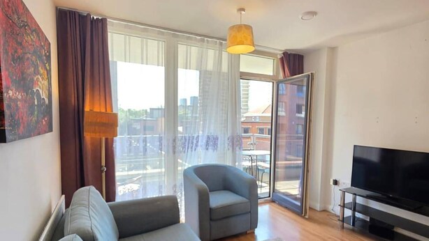A modern living room with a grey sofa, a small armchair, a flat-screen TV, and a large window with curtains. - Cozy City 1Br W/balcony at the Heart of Stratford (London)