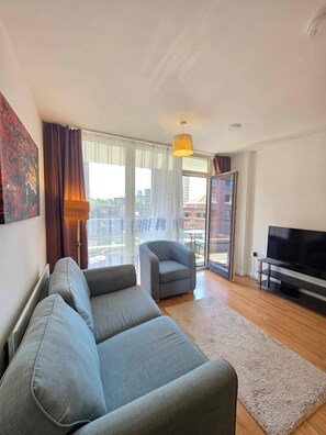 A modern living room with a grey sofa, a small armchair, a flat-screen TV, and a large window with curtains. - Cozy 1bdr With Balcony at the Heart of Stratford (London)