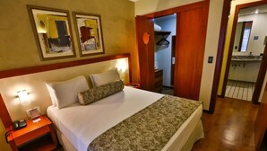 Habitación de hotel con cama, mesita de noche y espejo. Se ve una puerta que da a un baño y un pasillo.
