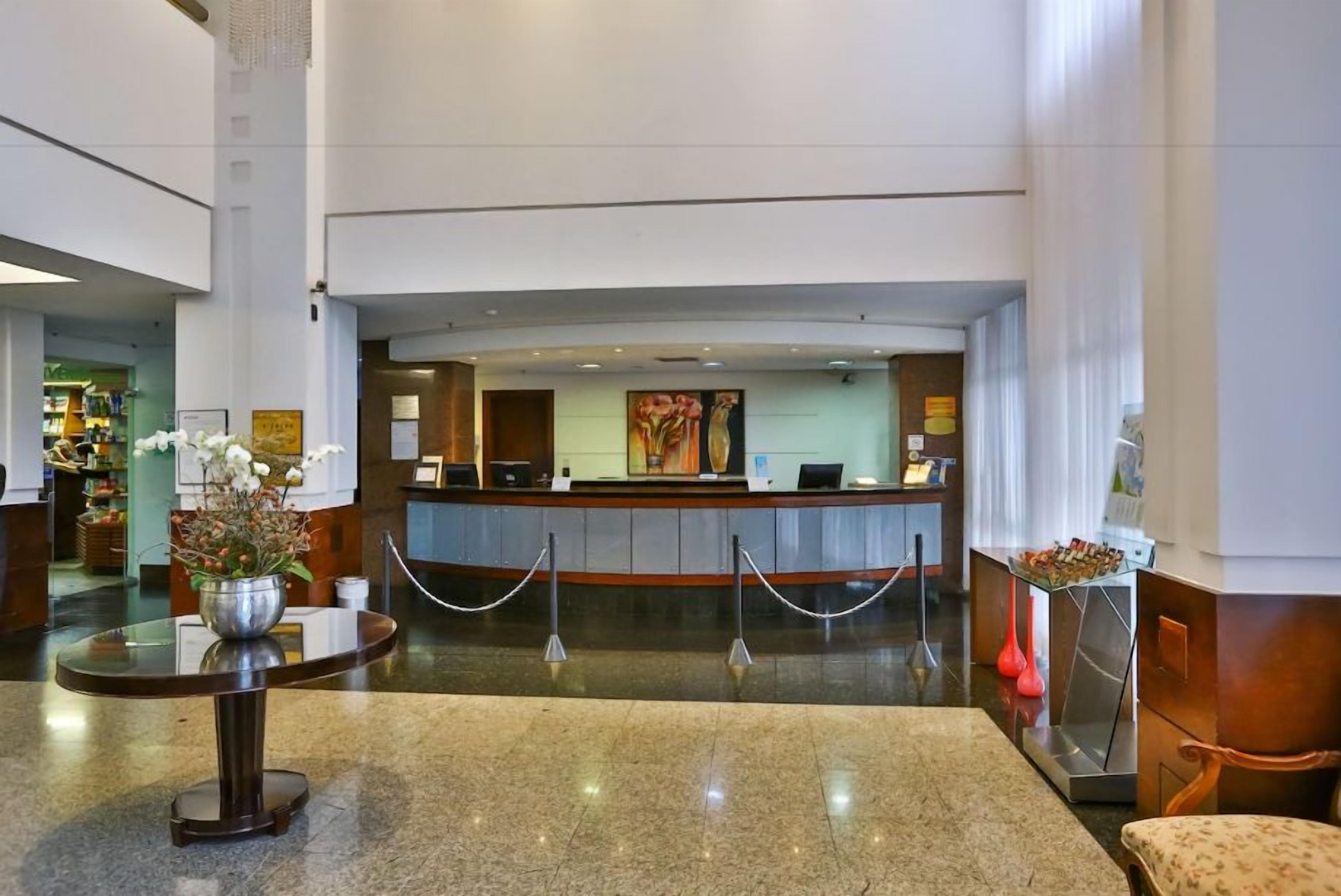 Zona de recepción de hotel con mostrador, área de espera con mesa y sillas, y un jarrón decorativo con flores.