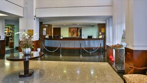 Zona de recepción de hotel con mostrador, área de espera con mesa y sillas, y un jarrón decorativo con flores.